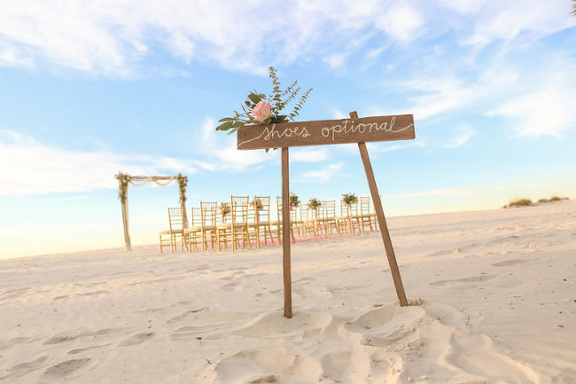 A beach wedding ceremony with a 'Shoes Optional' wooden sign