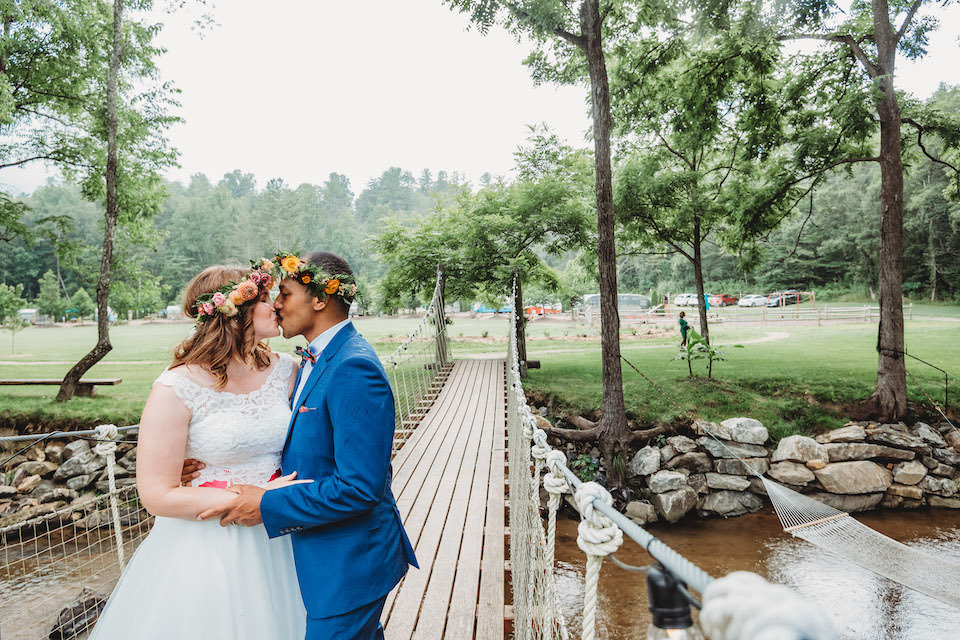 Newlyweds wearing floral crowns kiss in front of a bridge at JuneBug Retro Resort | 21 Hidden Wedding Costs