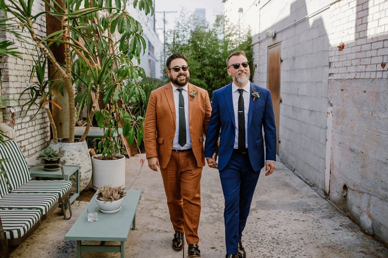 Two grooms in chic suits walk through the patio of the Grass Room | Hidden Wedding Costs To Budget For