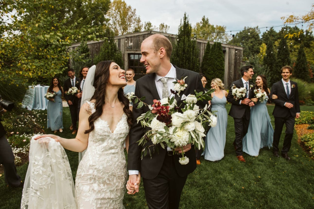 A bride and groom look at each other lovingly followed by their wedding party in front of The Field House barn